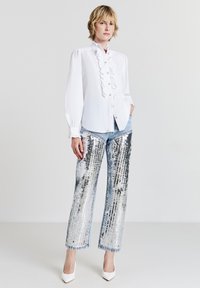 Blusa bianca con dettagli arricciati, maniche lunghe a sbuffo; abbinata a pantaloni larghi con paillettes argentate; modello che indossa tacchi bianchi.