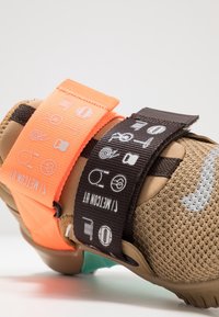 Brauner Sneaker mit Netzstruktur, ausgestattet mit einem orangefarbenen Riemen und einem schwarzen Riemen mit Icons. Auffällige Details sind die kontrastierenden Farben und Muster.