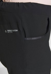 Svarta DuraStretch aktiva byxor med en texturerad finish, utrustad med ett elastiskt midjeband och en sidficka med en diskret logotyp.