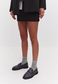 Zwarte leren loafers met een gladde afwerking, gecombineerd met lichtgrijze geribbelde sokken. Het model draagt een zwarte rok en blazer.