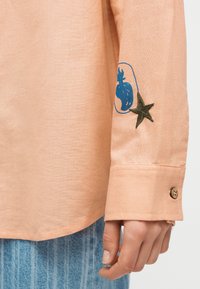 Κοντινό πλάνο του επωμίδας ενός μακρυμάνικου πουκάμισου ροδάκινου χρώματος με πράσινο κεντημένο αστέρι και μπλε αφηρημένο σχέδιο από πάνω, φορεμένο με ανοιχτό μπλε τζιν.