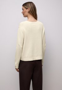 Pull de couleur crème à manches longues, avec poignets et ourlet côtelés, présenté avec un pantalon marron, doté d'une coupe décontractée et d'une texture douce.