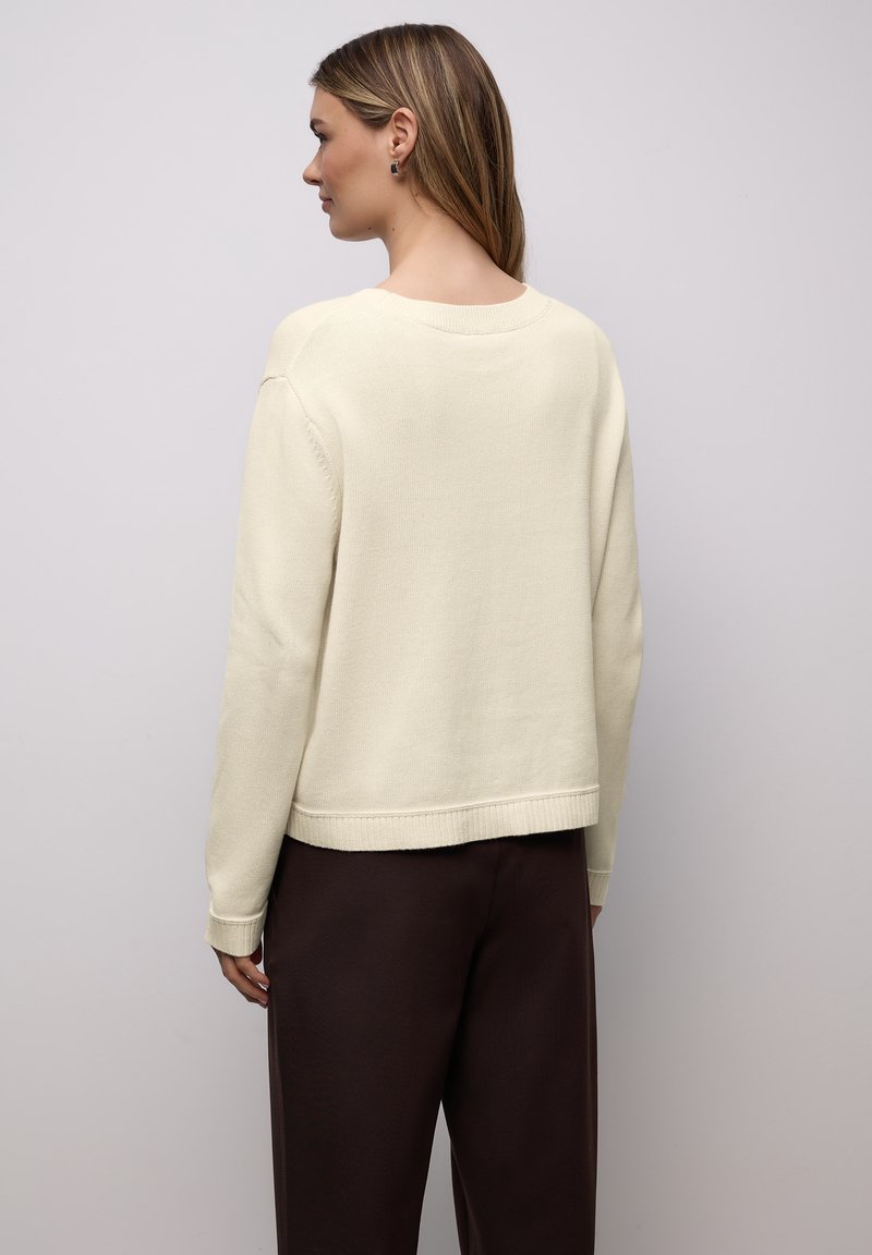 Pull de couleur crème à manches longues, avec poignets et ourlet côtelés, présenté avec un pantalon marron, doté d'une coupe décontractée et d'une texture douce.