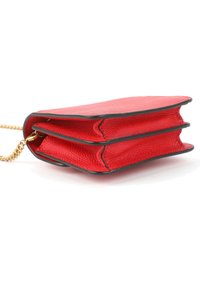 Pequeño bolso de cuero rojo con ribete negro y cadena dorada, mostrado de lado sobre un fondo blanco.
