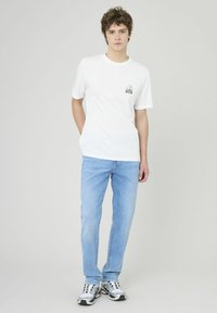 T-shirt en coton blanc avec un col rond et un petit motif multicolore sur le côté gauche de la poitrine, associé à un jean bleu clair et des baskets grises.