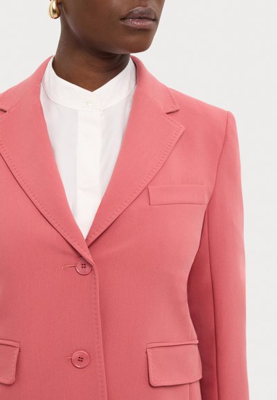 Pink blazer with notch lapels, two buttons, and front pockets. Fabric is smooth with a tailored fit, paired with a white collared top beneath.