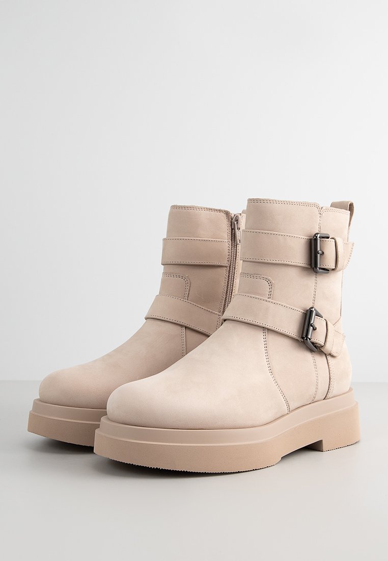 Beige Wildleder-Stiefeletten mit zwei verstellbaren Schnallen und einem seitlichen Reißverschluss, ausgestattet mit einer klobigen, beigen Plattformsohle und einer glatten Textur.