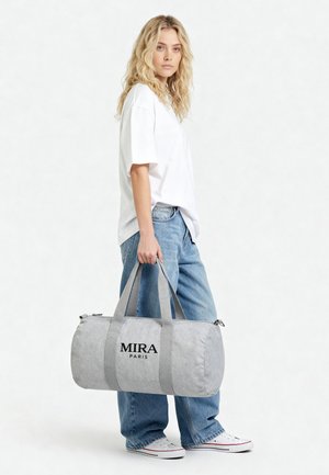 Jeune femme en t-shirt blanc et jean bleu tenant un sac de sport cylindrique gris clair avec le logo "MIRA PARIS".