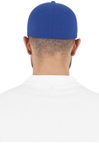 Căciulă de baseball albastră cu cusături, coroană rotundă și spate ajustabil, purtată de o persoană în cămașă polo albă, văzută din spate.