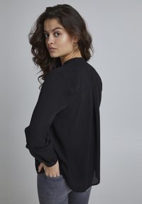 Zwarte lange mouwen blouse met een gladde textuur, voorzien van een plooitje aan de achterkant en knoopmanchetten. Model draagt een grijze jeans.