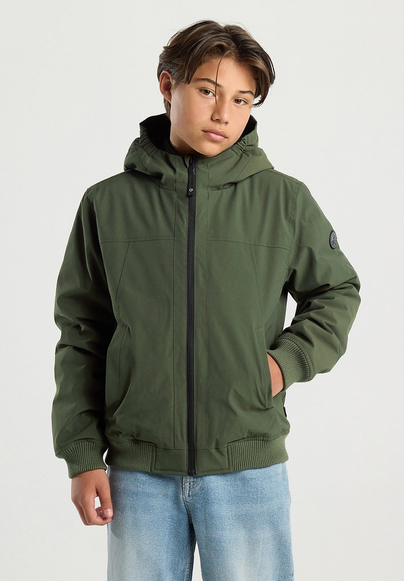 Teenager with short brown hair wearing an olive green hooded jacket and light blue jeans, standing with one hand in pocket.