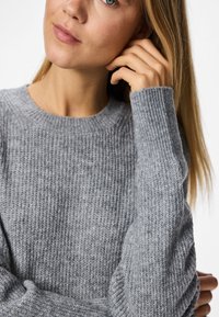 Γκρι ριχτό πουλόβερ με στρογγυλή λαιμόκοψη και μακριά μανίκια, φτιαγμένο από μαλακό, υφασματώδη ύφασμα, δείχνοντας μια κοντινή άποψη του πλέγματος.