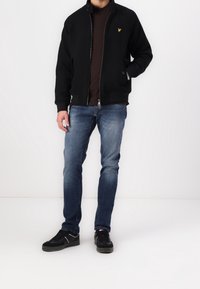 Schwarze Bomberjacke mit Reißverschluss und gerippten Bündchen, getragen über einem dunklen Hemd, kombiniert mit blauen Jeans und schwarzen Sneakers mit weißen Akzenten.