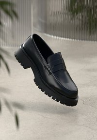 Black leather loafers with a raised sole, featuring a decorative strap across the vamp. Smooth texture and rounded toe design.