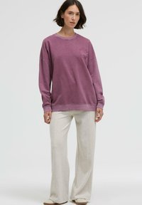 Lila Baumwollpullover mit langen Ärmeln und Rundhalsausschnitt, mit einem runden Logo-Print. Kombiniert mit hellbeigen weiten Hosen.