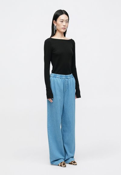 Melnas garās piedurknes blūze, gaiši zila plati kāju džinsi ar elastīgu jostasvietu un šauras leopardu raksta kurpes. Modelis stāv profilā.