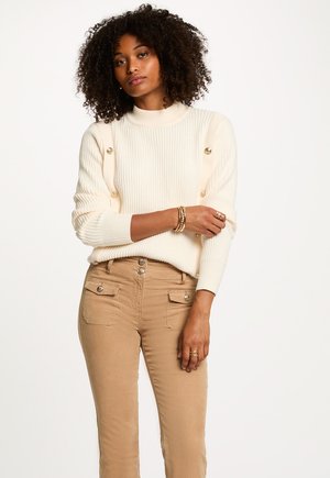 Pull en côtes crème avec des accents de boutons dorés aux épaules, associé à un pantalon taille haute beige avec des poches à boutons.