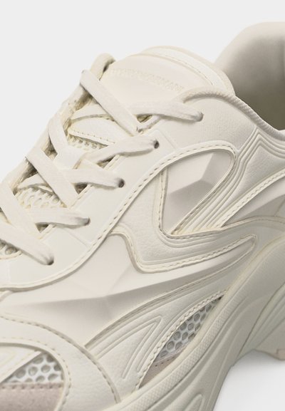 Baskets couleur crème en cuir texturé, panneaux en maille, coutures complexes et lacets blancs, vue rapprochée du côté supérieur.