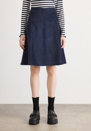 Femme portant une jupe trapèze bleu marine avec des poches latérales, associée à des chaussettes noires, des chaussures à boucles noires et un haut à manches longues rayé noir et blanc.