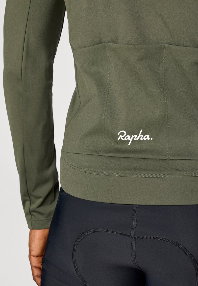 Olivgrön cykeljacka med långa ärmar, med en mjuk textur och "Rapha"-logotyp i vitt. Svarta cykelshorts med vadderade detaljer.