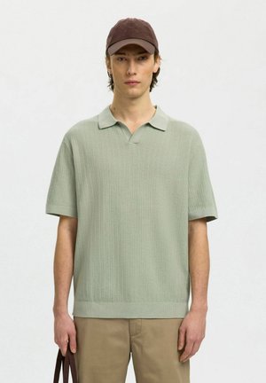 Jeune homme portant un polo à manches courtes texturé vert clair, un pantalon beige et une casquette de baseball marron, tenant la poignée d'un sac marron foncé.