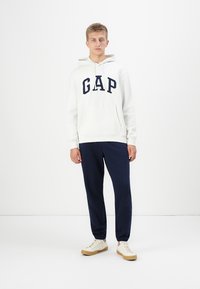 Hvit hettegenser med marineblå "GAP" bokstaver, kengurulomme, kombinert med marineblå joggebukser og lyse sneakers. Enkelt design, uformell stil.