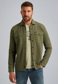 Olijfgroene geweven shirtjack met twee borstzakken, knoopsluiting en een gestructureerde stof; gecombineerd met een lichtgroene T-shirt en jeans.