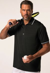 Polo noir avec des accents blancs, doté d'une courte fermeture éclair et d'un logo. Le modèle tient une raquette de tennis et une balle blanche.