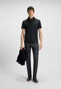 Mørk polo skjorte med en blød tekstur, parret med smalle, mørkegrå bukser. Modellen holder en marineblå jakke. Afslappet fodtøj fuldender looket.