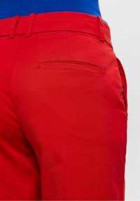 Gros plan sur un pantalon rouge avec poche passepoilée à l'arrière et passants de ceinture, porté avec une chemise bleue rentrée, sur un fond clair.