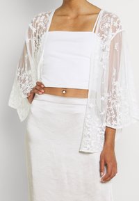 Kvinde iført hvid blonde kimono over hvidt crop top og cremefarvet nederdel med en navlepiercing, stående med den ene hånd på hoften.
