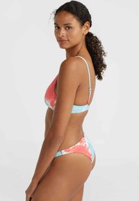 Kvinna med lockigt hår i en blå och korallfärgad tie-dye bikini, vänd åt sidan och ser över axeln mot en enfärgad bakgrund.