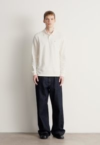 Balta polo krekls ar garām piedurknēm kombinēts ar tumšām platajiem džinsiem. Modelis stāv ar kājām kopā, valkājot melnus apavus. Vienkāršs fons.