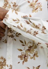 Bílá květovaná košile s hnědými a zlatými květinovými vzory. Má límec a manžety na knoflíky, vyrobená z hladké látky.