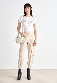 Beige Cargo-Hosen mit elastischen Bündchen, zwei Seitentaschen und einer taillierten Taille. Das weiße T-Shirt hat ein grafisches Design in Grau. Schwarze Ankle Boots.