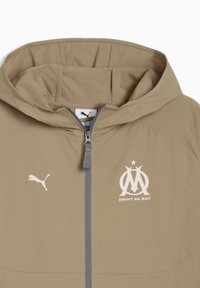 Μπεζ μπουφάν με κουκούλα, με φερμουάρ μπροστά και λογότυπο με το κείμενο "DROIT AU BUT." Περιλαμβάνει το λογότυπο της Puma στη αριστερή πλευρά. Με λεία υφή.
