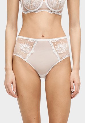 Femme portant un sous-vêtement beige taille haute avec des panneaux en dentelle florale et un soutien-gorge assorti en dentelle, debout devant un fond uni.