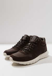 Paire de baskets montantes marron foncé avec semelles blanches texturées mouchetées et lacets noirs sur une surface blanche.
