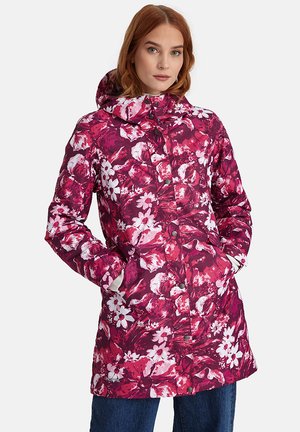 Naine kannab põlveni ulatuvat kapuutsiga jopet roosa, punase ja valge lillemustriga, käed taskutes, kombineeritud siniste teksadega.