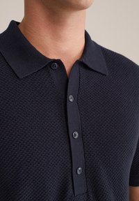 Tuvumā redzams cilvēks, kurš valkā tumši zilu teksturētu polo kreklu ar apkakli un četriem pogām uz apģērba priekšpuses.