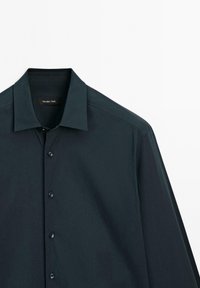 Tmavě zelená košile s límcem na knoflíky, má hladkou texturu, dlouhé rukávy a šest odpovídajících knoflíků vpředu. Viditelná značka.