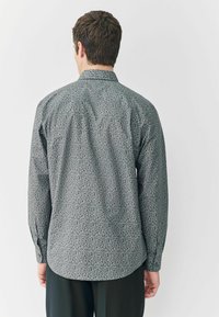 Hemd mit Knöpfen aus einem dunkelgrünen Stoff mit kleinem Blumenmuster. Verfügt über lange Ärmel, einen traditionellen Kragen und einen abgerundeten Saum.