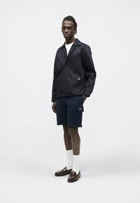 Chaqueta de algodón azul marino con un bolsillo en el pecho y dos bolsillos delanteros, combinada con pantalones cortos azul marino con un bolsillo lateral y calcetines blancos hasta la mitad de la pantorrilla.