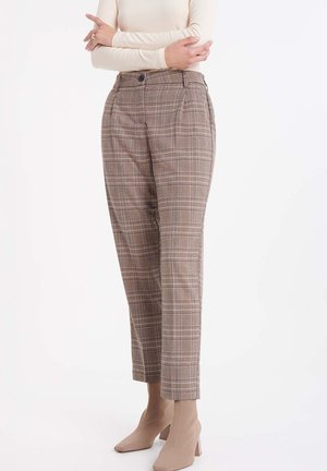 Vrouw draagt bruine geruite broek met hoge taille, gecombineerd met beige sokstijl enkellaarsjes met hak, op een witte achtergrond.