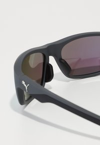 Lunettes de sport avec une monture noir mat et des verres teintés violets. Comprend des plaquettes de nez en caoutchouc et un accent logo argenté sur le côté.