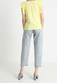 Žlutý top s volánovými rukávy a přiléhavým střihem, v kombinaci s světle modrými rovnými džínami. Modelka má na sobě černobílé pruhované vysoké boty.