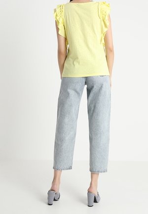 Geel rimpel mouwtop met een getailleerd ontwerp, gecombineerd met lichtblauwe rechte jeans. Model draagt zwart-witte gestreepte hakschoenen.