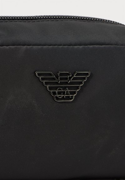 Must musta kott musta tekstiiliga, millel on metallikmust logo tiibadega ja keskmes "GA", ning tõmblukk.