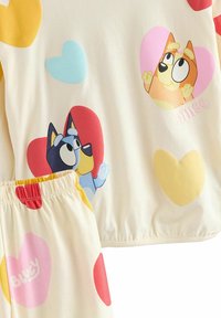 Set di pigiama a due pezzi color crema chiaro con motivi di cuori colorati. Include immagini dei personaggi dei cartoni animati Bluey e Bingo in vari colori.