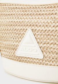 Sac en paille tressée avec une surface texturée beige, présentant un large patch triangulaire blanc avec "ALDO" en relief au centre.